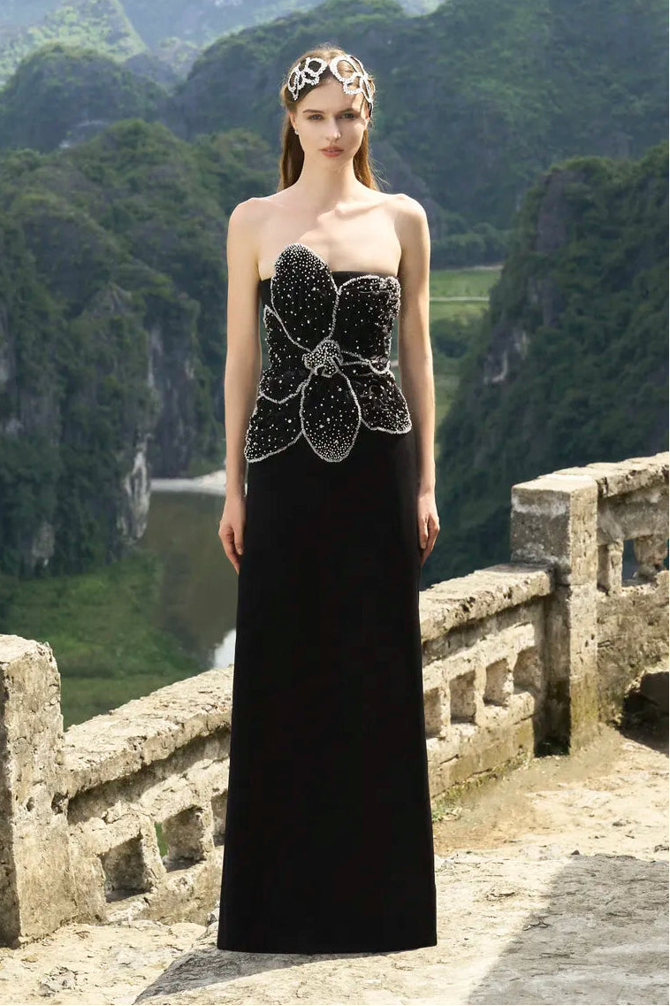 Woman in a black evening gown with a floral embellishment standing on a stone platform with mountains in the background.